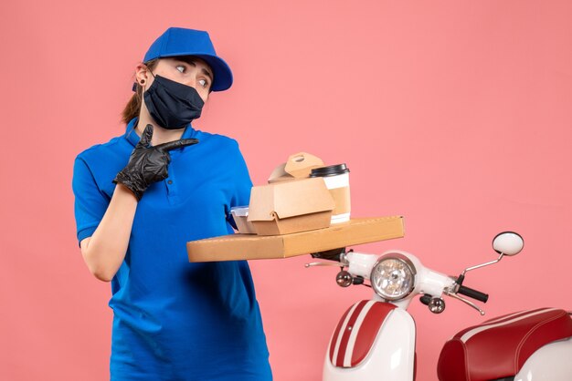 Вид спереди женщина-курьер в маске с доставкой кофе и еды на розовом полу пандемия covid - доставка униформы работника службы занятости