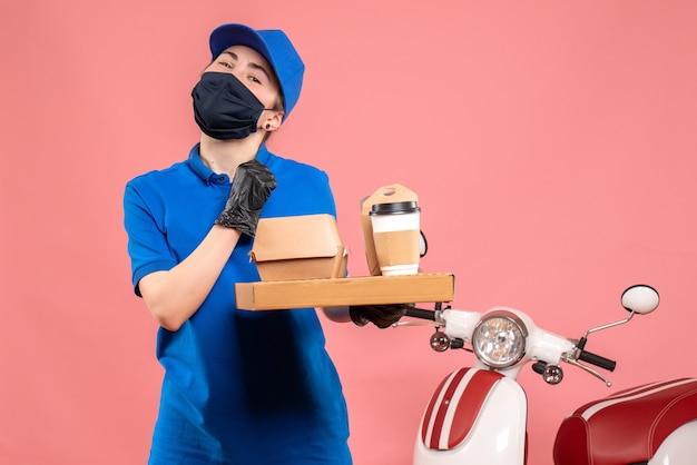 Front view female courier in mask with delivery coffee and food on a pink 