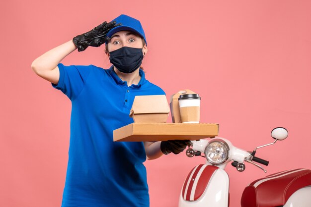 Front view female courier in mask with delivery coffee and food on pink 