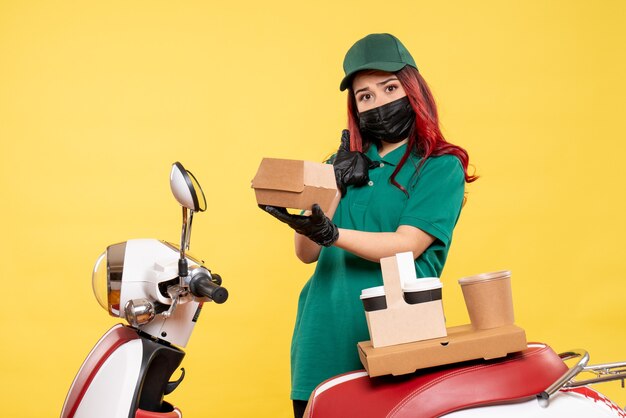 Front view of female courier in mask with delivery coffee and food on the yellow wall