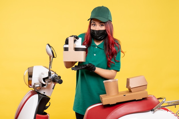 Front view of female courier in mask with delivery coffee and food on yellow wall