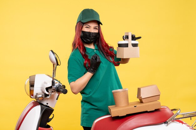 Front view of female courier in mask with delivery coffee and food on yellow wall