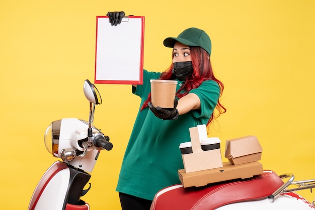 Front view of female courier in mask with delivery coffee and food on yellow wall