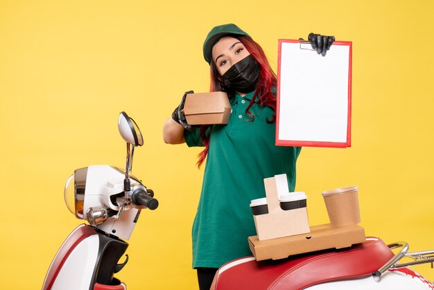 Front view of female courier in mask with delivery coffee and food on yellow wall