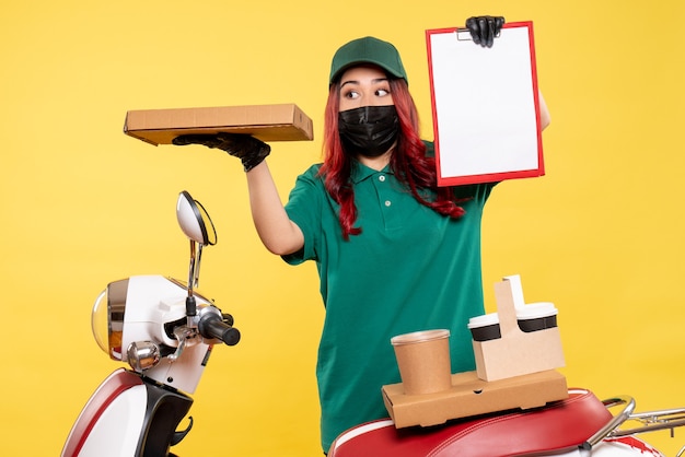 Front view of female courier in mask with delivery coffee and food on yellow wall