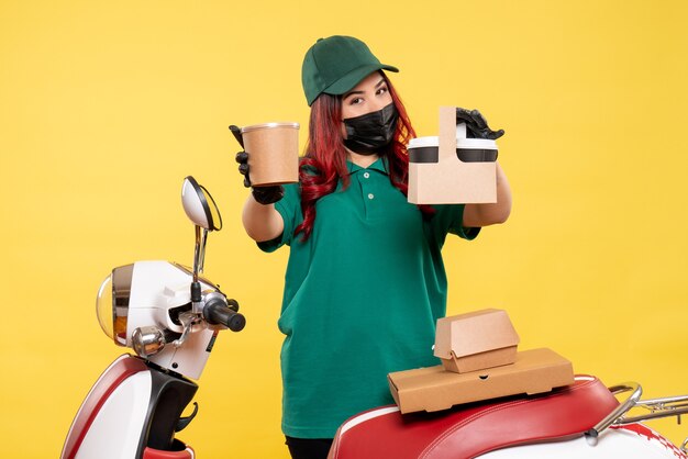 Front view of female courier in mask with delivery coffee on a yellow wall