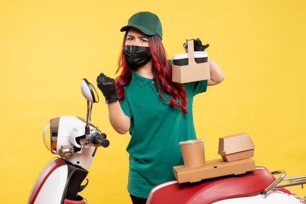 Front view of female courier in mask with delivery coffee on yellow wall