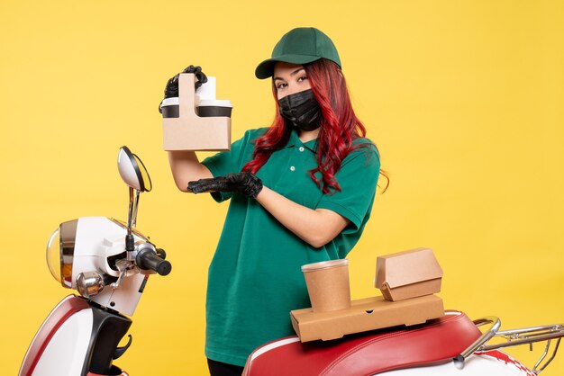 Front view of female courier in mask with delivery coffee on yellow wall