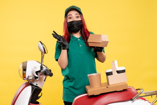 Front view of female courier in mask with delivery coffee on yellow wall