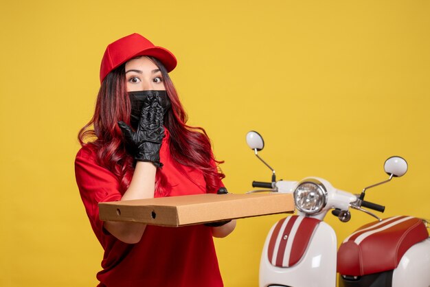 Front view of female courier in mask with delivery food box on a yellow wall