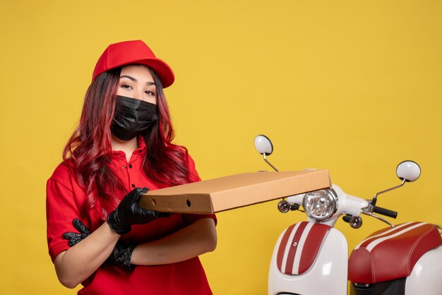 Front view of female courier in mask with delivery food box on the yellow wall