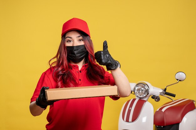 Front view of female courier in mask with delivery food box on yellow wall