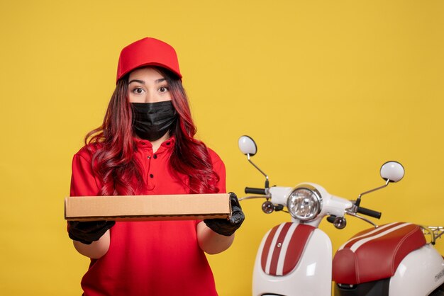 Front view of female courier in mask with delivery food box on yellow wall