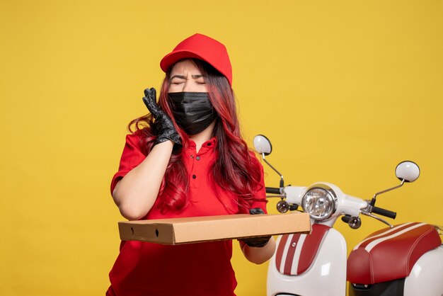 Front view of female courier in mask with delivery food box on yellow wall