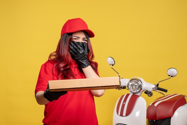 Front view of female courier in mask with delivery food box on yellow wall