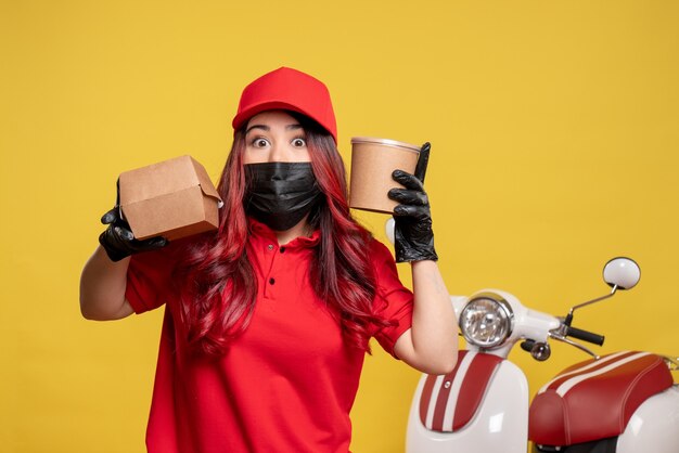 Front view of female courier in mask with delivery food and dessert on yellow wall