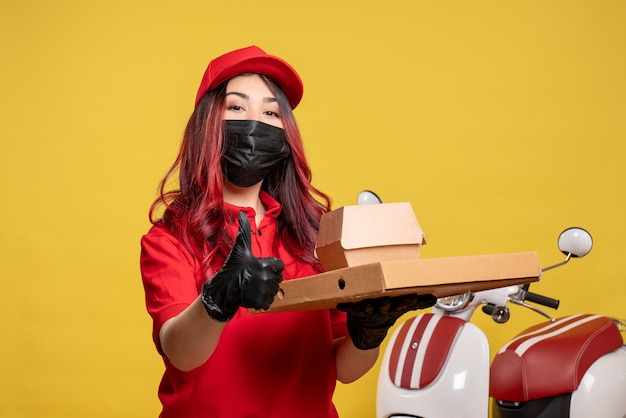 Front view of female courier in mask with delivery food on a yellow wall