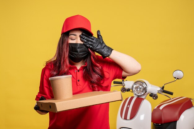 Front view of female courier in mask with delivery food on the yellow wall