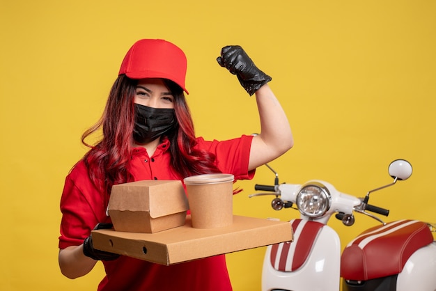 Front view of female courier in mask with delivery food on yellow wall