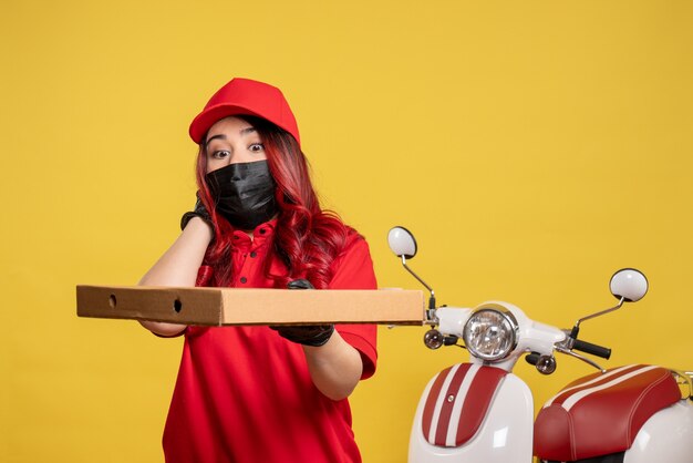 Front view of female courier in mask with delivery food on yellow wall