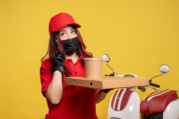 Front view of female courier in mask with delivery food on yellow wall