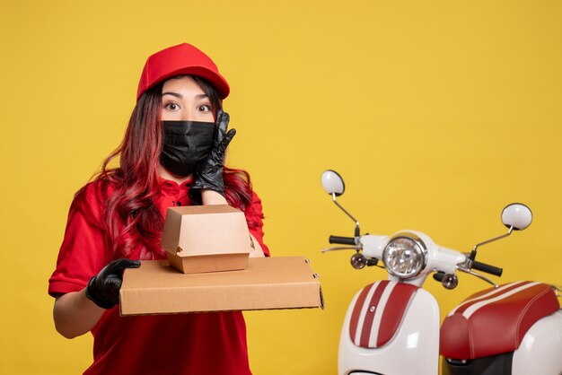 Front view of female courier in mask with delivery food on yellow wall