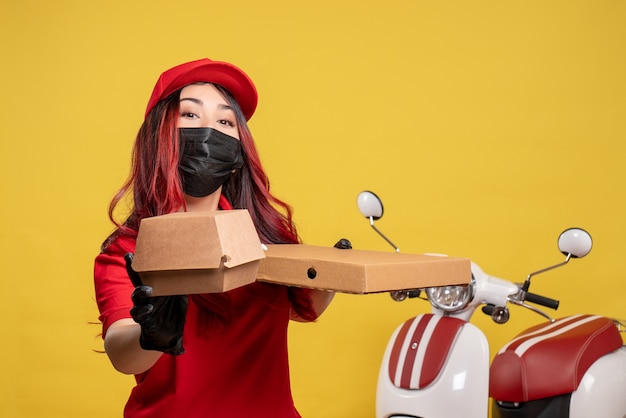 Front view of female courier in mask with delivery food on yellow wall