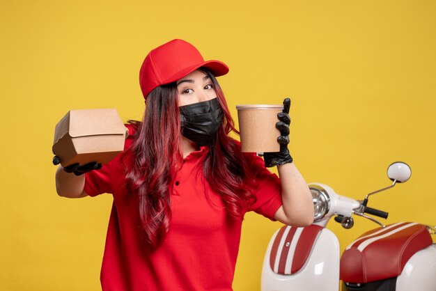 Front view of female courier in mask with delivery food on yellow wall