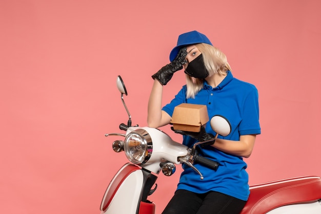 Front view female courier in mask with little food package on a pink 