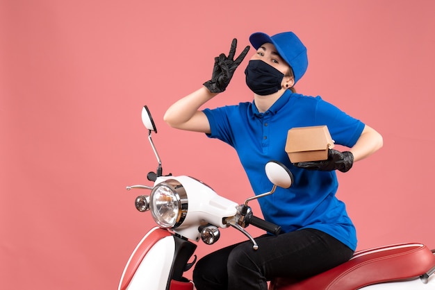 Front view female courier in mask with little food package posing on pink 