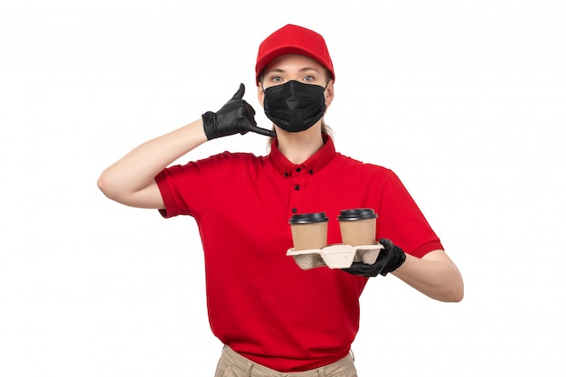 A front view female courier in red shirt red cap black gloves and black mask holding coffee asking to call posing on white