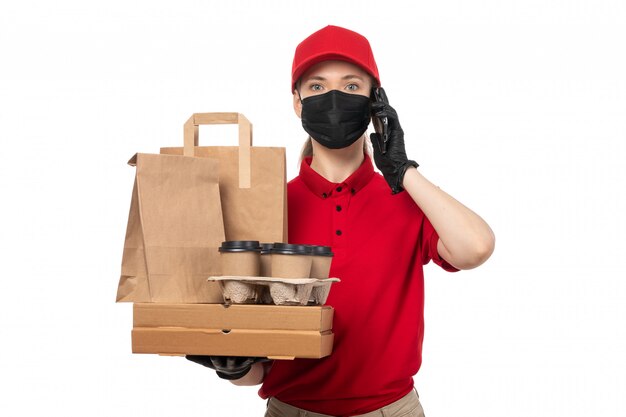 A front view female courier in red shirt red cap black gloves and black mask holding pizza boxes and coffee cups on white