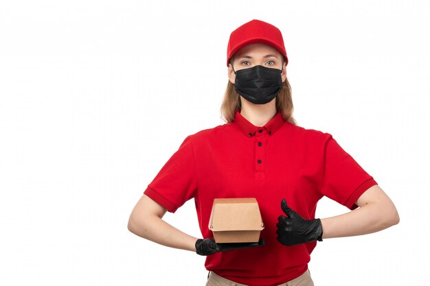 A front view female courier in red shirt red cap black gloves and mask holding package with food on white