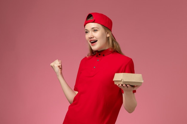 Front view female courier in red uniform and cape cheering holding little delivery food package on pink wall, delivery service company uniform job