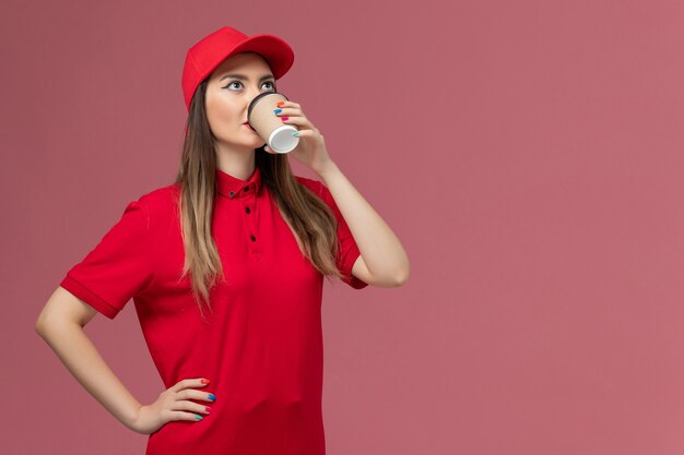 Вид спереди женщина-курьер в красной форме и накидке, пьющая кофе на розовом фоне, форма доставки службы, рабочий