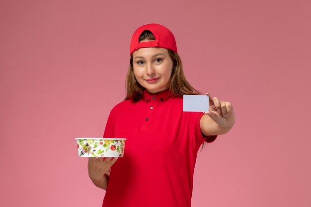 Front view female courier in red uniform and cape holding delivery bowl and card on pink wall, uniform delivery service job worker