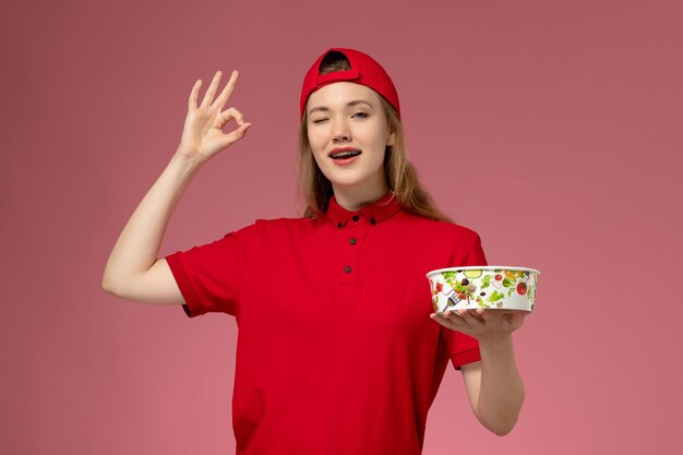 Front view female courier in red uniform and cape holding delivery bowl on light-pink wall, work service uniform delivery