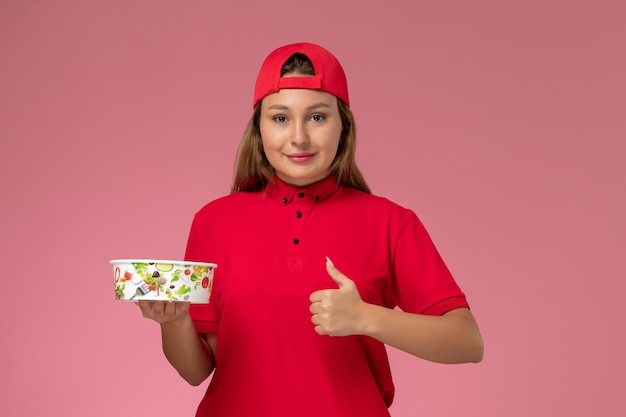 Front view female courier in red uniform and cape holding delivery bowl on pink wall, uniform delivery service job