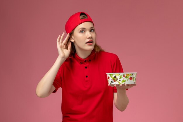 Front view female courier in red uniform and cape holding delivery bowl trying to hear on the light pink wall, service uniform delivery worker