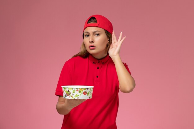 빨간색 유니폼과 케이프 핑크 벽, 균일 한 배달 서비스에 들으려고 배달 그릇을 들고 전면보기 여성 택배