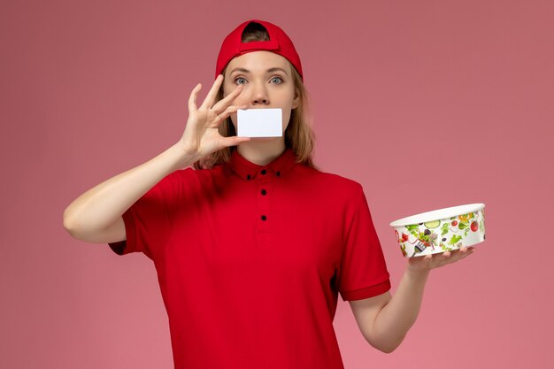 Front view female courier in red uniform and cape holding delivery bowl with card on light pink wall, job service uniform delivery