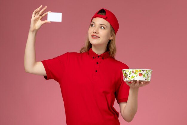 Front view female courier in red uniform and cape holding delivery bowl with card on light pink wall, worker service uniform delivery