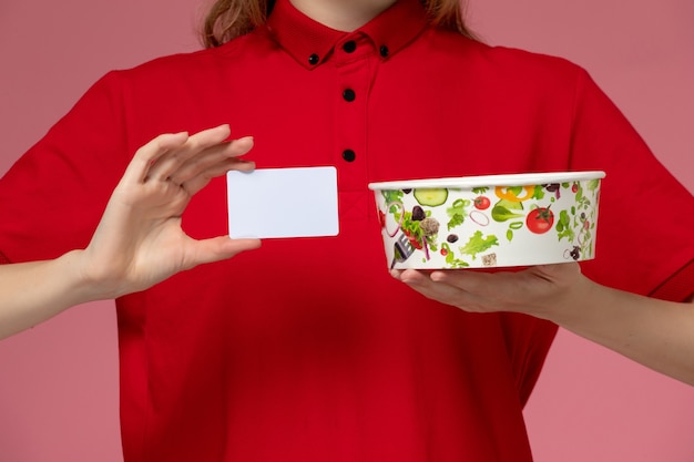 Front view female courier in red uniform and cape holding delivery bowl with card on the light-pink wall, worker service uniform delivery