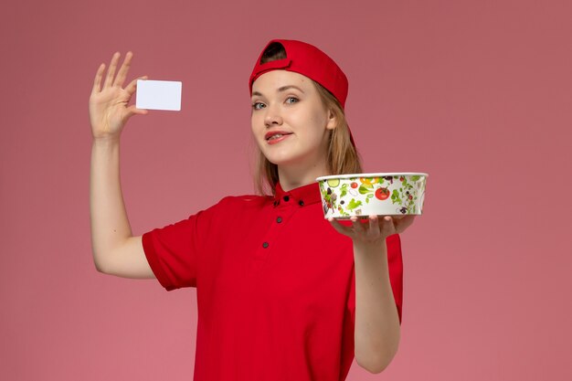 赤い制服を着た正面図の女性の宅配便と淡いピンクの壁に笑顔のカードと配達ボウルを保持している岬、労働者サービスの制服配達