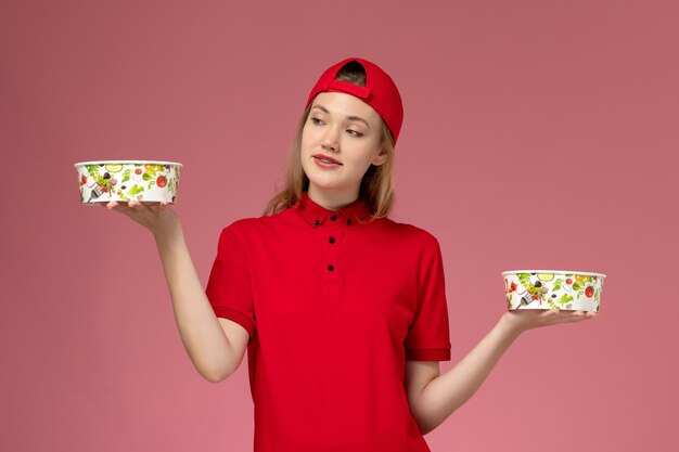 Front view female courier in red uniform and cape holding delivery bowls on the light-pink wall, service job uniform delivery