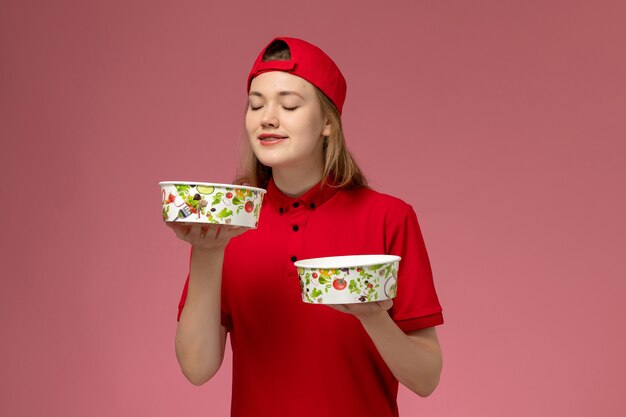 Front view female courier in red uniform and cape holding delivery bowls on light pink wall, service job worker uniform delivery