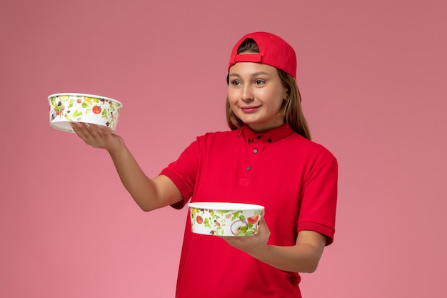 Front view female courier in red uniform and cape holding delivery bowls on pink wall, uniform delivery job service