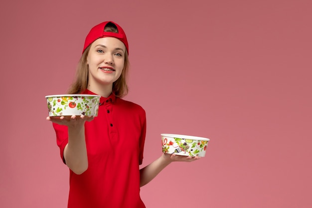 Вид спереди женщина-курьер в красной форме и накидке, держащая миски для доставки с улыбкой на светло-розовом столе, доставка униформы