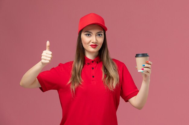 Вид спереди женщина-курьер в красной форме и плаще, держащая чашку кофе на светло-розовом фоне