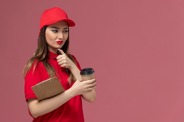 Corriere femminile di vista frontale in uniforme rossa e mantello che tiene il blocco note e la penna della tazza di caffè di consegna su fondo rosa-chiaro dell'uniforme di consegna del servizio